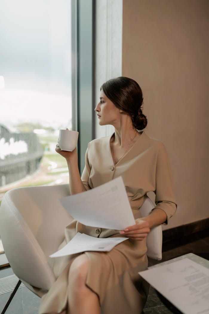 Sophisticated woman in beige dress holding a cup of coffee and papers, seated indoors beside a window.